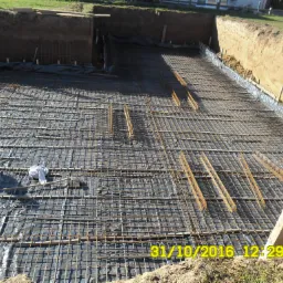 Przygotowanie zbrojenia na gruncie pokrytym folią, widoczne wykopy ziemne i wystające pręty zbrojeniowe.