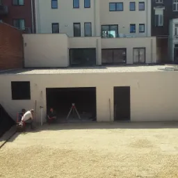 Przygotowanie podłoża z warstwą tłucznia przed budynkiem z garażem; pracownicy z niwelatorem laserowym i ubijarką płytową.