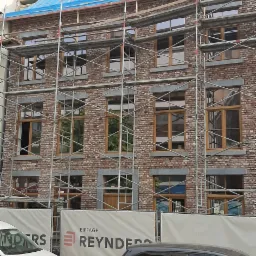 Wielopiętrowy budynek z cegły na etapie budowy, z widocznymi rusztowaniami, oknami z drewnianymi ramami i niebieską folią ochronną na dachu, z trzema lukarnami w trakcie montażu, z banerem...