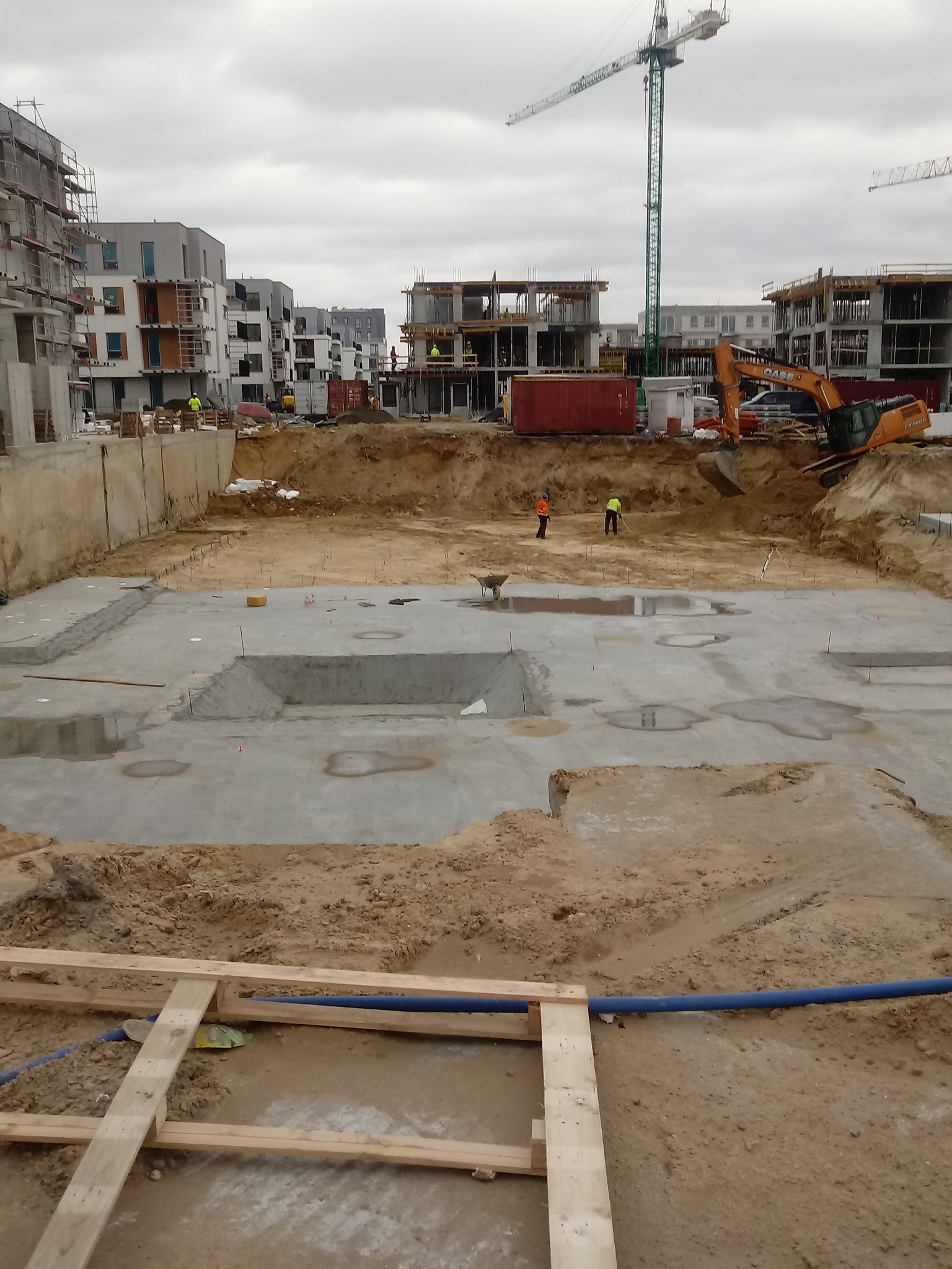 Wykop pod fundamenty nowego osiedla z widoczną wylaną płytą fundamentową, pracownikami w odblaskowych kamizelkach, koparką i dźwigiem w tle.