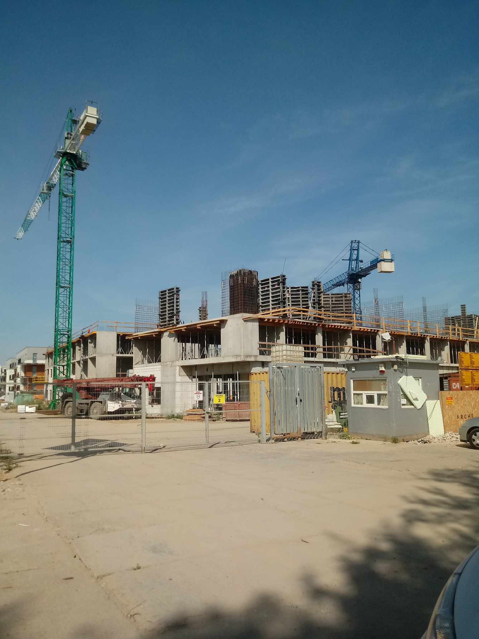 Wznoszenie budynku z żelbetu, widoczne szkielety zbrojeniowe, szalunki i dwa żurawie wieżowe na tle błękitnego nieba.