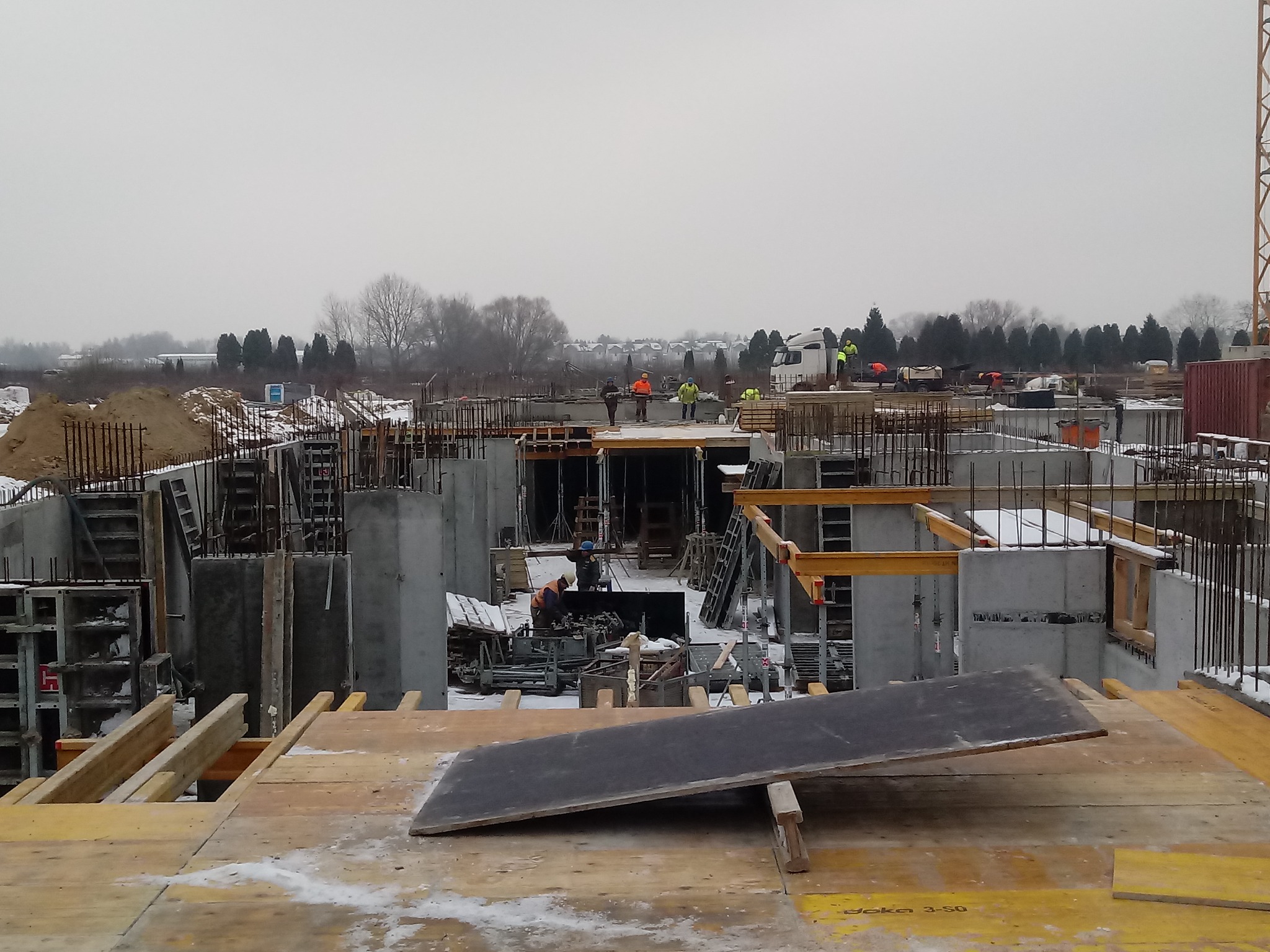 Szeroki plan budowy z betonu w szarej zimowej scenerii, widoczne zbrojenia, szalunki i pracujący robotnicy w pomarańczowych kamizelkach. W tle drzewa i niebo pokryte chmurami.
