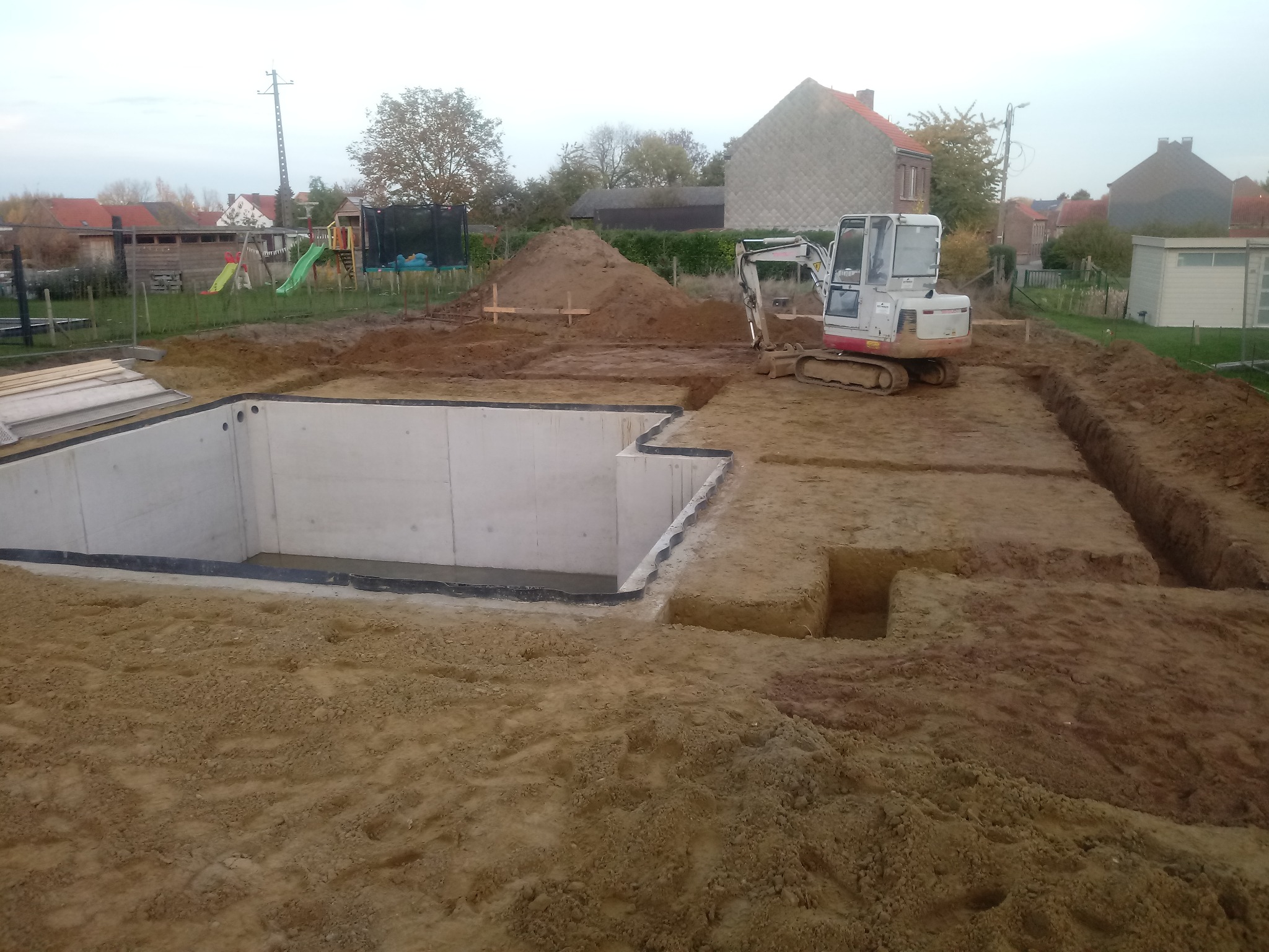 Budowa fundamentów pod basen: wykop z betonowymi ścianami, mała koparka i sterta ziemi na działce z placem zabaw w tle.