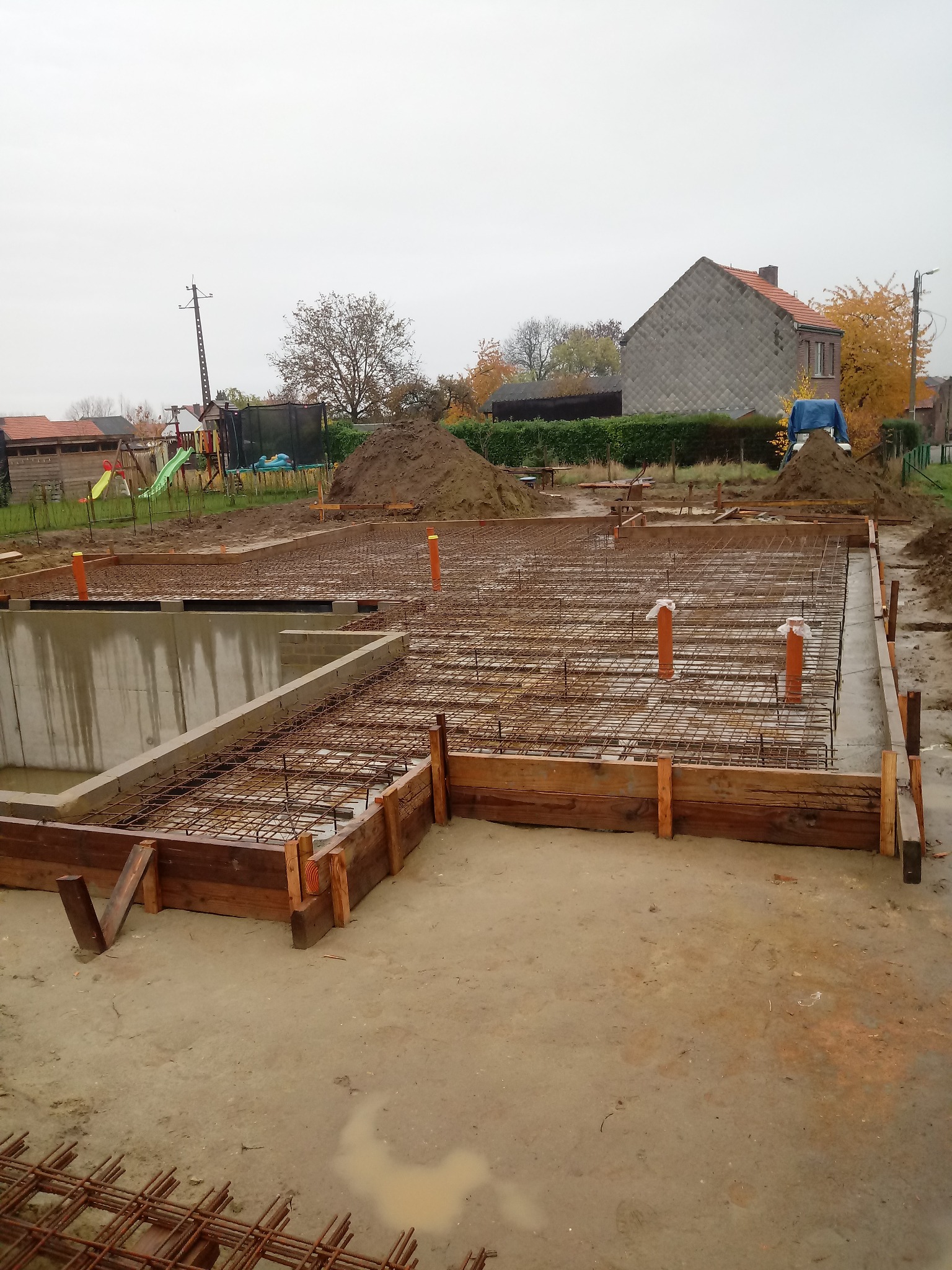 Przygotowany szalunek i zbrojenie pod płytę fundamentową domu jednorodzinnego, widoczne pionowe rury instalacyjne, w tle zabudowa jednorodzinna i plac zabaw.