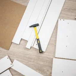 Rozłożone białe elementy meblowe, płyta wiórowa i młotek z żółtą rękojeścią na podłodze z paneli, przygotowane do montażu.