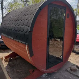 Gotowa beczka sauny z drewnianą obudową w kolorze czerwonym i czarnym, z widoczną szklaną szybą w drzwiach i kominem na dachu, ustawiona na betonowym podłożu.