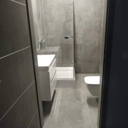 Kalmia Enterprise Sp zoo - Nowoczesna łazienka z szarymi płytkami, kabiną prysznicową ze szklaną ścianką, wiszącą białą umywalką z szafką i toaletą, widziana zza częściowo otwartych drzwi; fioletowy dywanik na podłodze.