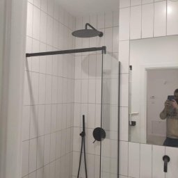 Kalmia Enterprise Sp zoo - Nowoczesna łazienka z kabiną prysznicową typu walk-in, czarną armaturą i białymi, wąskimi płytkami ułożonymi pionowo, odbicie osoby robiącej zdjęcie w lustrze.