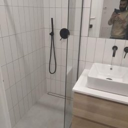 Kalmia Enterprise Sp zoo - Nowoczesna łazienka z kabiną prysznicową typu walk-in, białe pionowe płytki, czarna armatura i umywalka nablatowa na drewnianej szafce. W lustrze odbija się osoba robiąca zdjęcie.