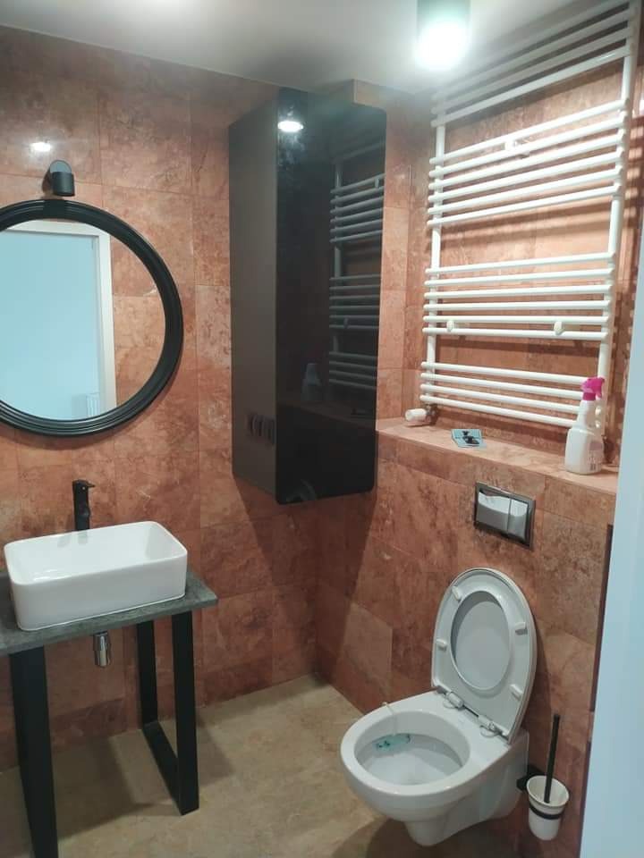 Wykończona łazienka z marmurowymi płytkami, wiszącą szafką z lustrzanym frontem, grzejnikiem drabinkowym, prostokątną umywalką nablatową na metalowym stelażu i otwartą toaletą z niebieskim płynem...