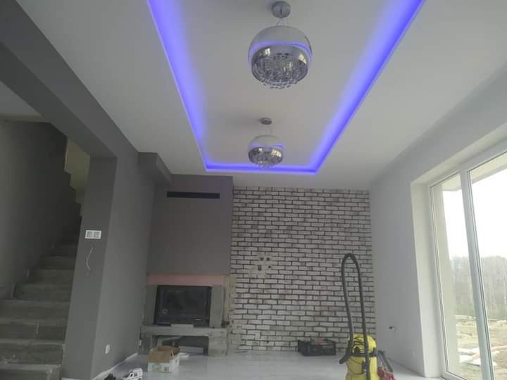 Wnętrze pokoju z kominkiem obłożonym białą cegłą, sufit z wnęką oświetloną niebieskim światłem LED, dwie lampy wiszące, schody w szarej tonacji po lewej stronie, odkurzacz na podłodze.