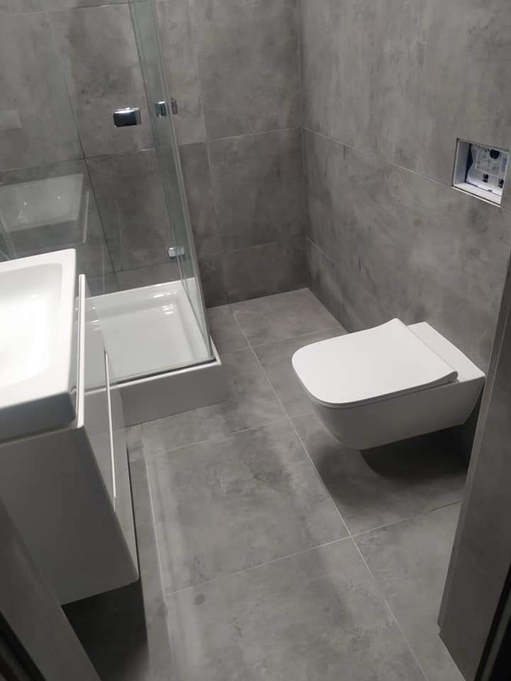 Nowoczesna łazienka z szarymi płytkami, wiszącą toaletą i kabiną prysznicową z przezroczystego szkła, widoczny fragment umywalki po lewej stronie.