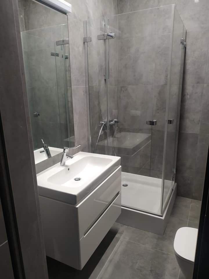 Nowoczesna łazienka w odcieniach szarości z białą umywalką na szafce i kabiną prysznicową ze szklanymi ściankami, widoczny fragment toalety.