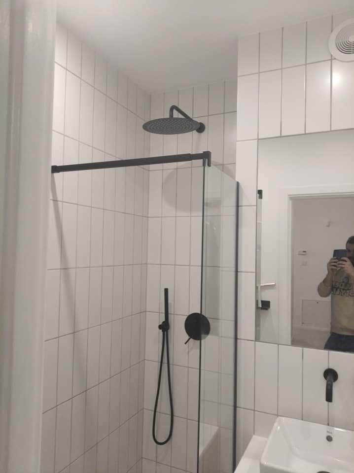 Nowoczesna łazienka z kabiną prysznicową typu walk-in, czarną armaturą i białymi, wąskimi płytkami ułożonymi pionowo, odbicie osoby robiącej zdjęcie w lustrze.