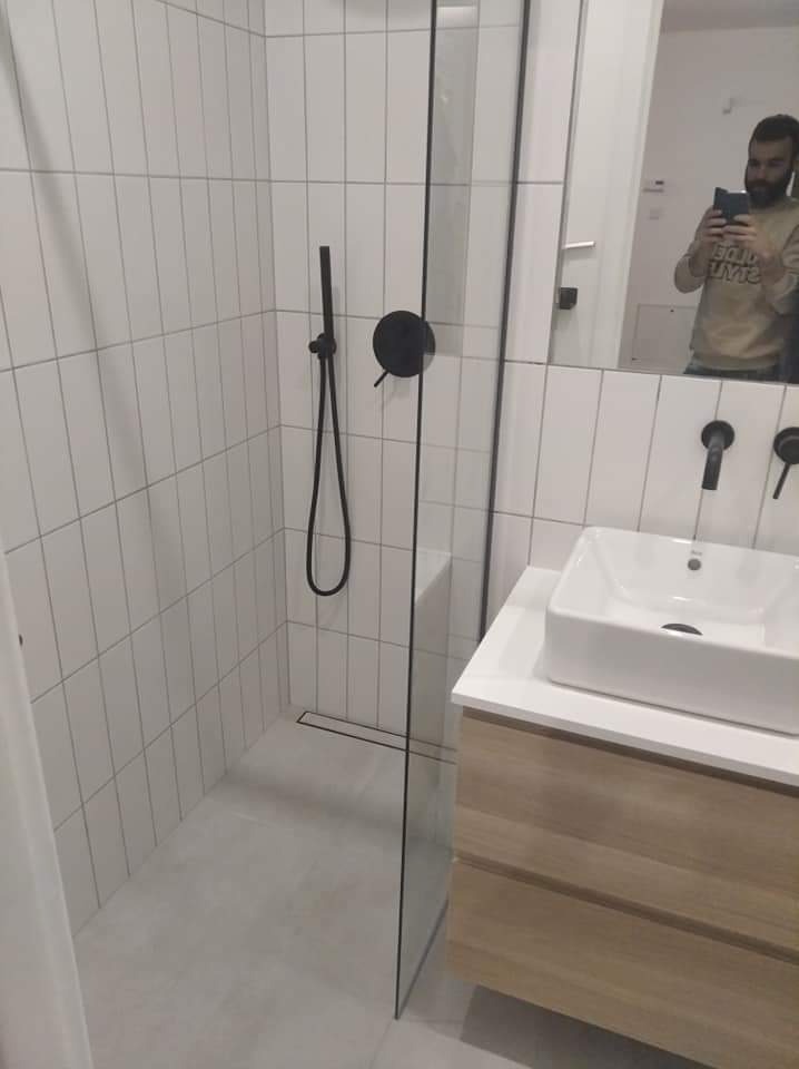 Nowoczesna łazienka z kabiną prysznicową typu walk-in, białe pionowe płytki, czarna armatura i umywalka nablatowa na drewnianej szafce. W lustrze odbija się osoba robiąca zdjęcie.