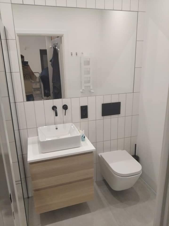 Nowoczesna łazienka z umywalką nablatową na drewnianej szafce, wiszącą toaletą i białymi płytkami ułożonymi pionowo, z czarną armaturą i osprzętem.