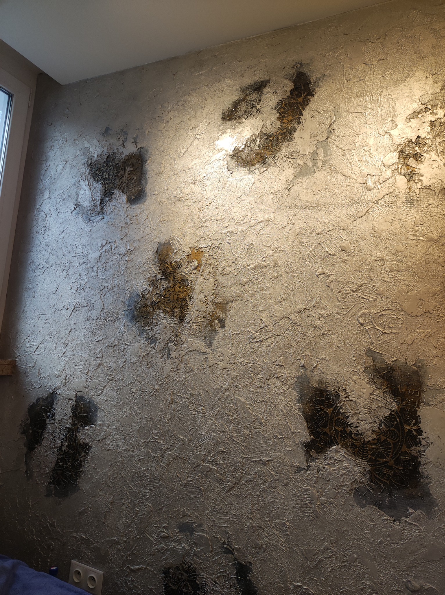 Dekoracyjna ściana z fakturą i ciemnymi, okrągłymi wzorami przypominającymi mandale, oświetlona od góry, z widocznym gniazdkiem elektrycznym w dolnej części.