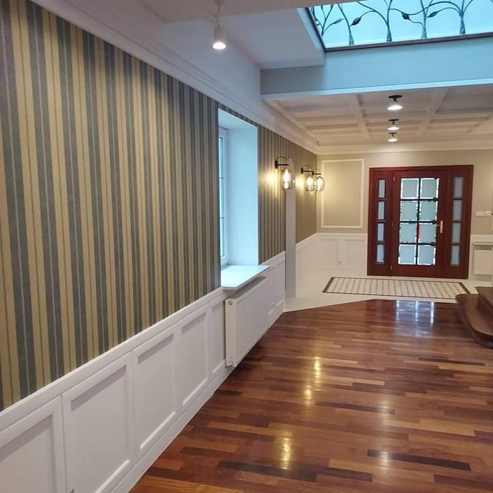 Korytarz po remoncie z tapetą w pionowe pasy, drewnianą podłogą i dekoracyjnym oknem dachowym z motywem roślinnym.