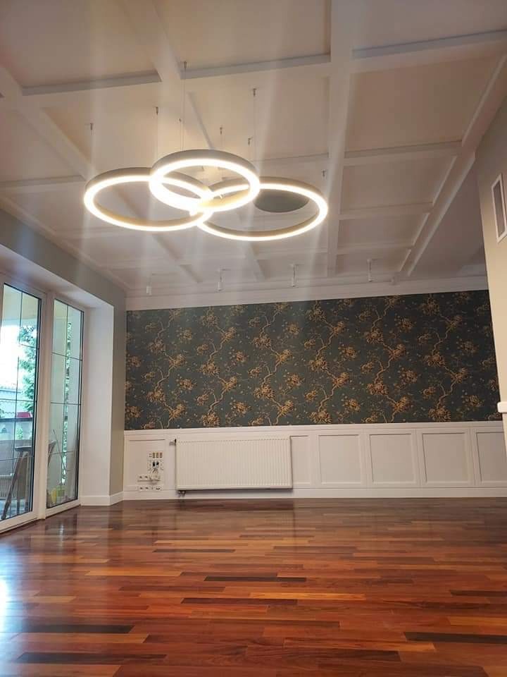 Salon z błyszczącą drewnianą podłogą, ozdobną tapetą w kwiaty, białymi panelami ściennymi i nowoczesną lampą sufitową z trzema świetlistymi okręgami.