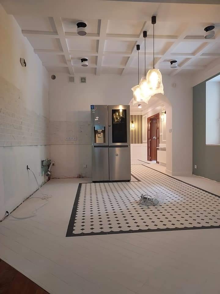 Wnętrze w trakcie prac wykończeniowych: białe drewniane podłogi, fragment mozaiki podłogowej, ściana z surowej cegły i nowoczesna lodówka z czarnym panelem, zwisające lampy z mlecznego szkła...