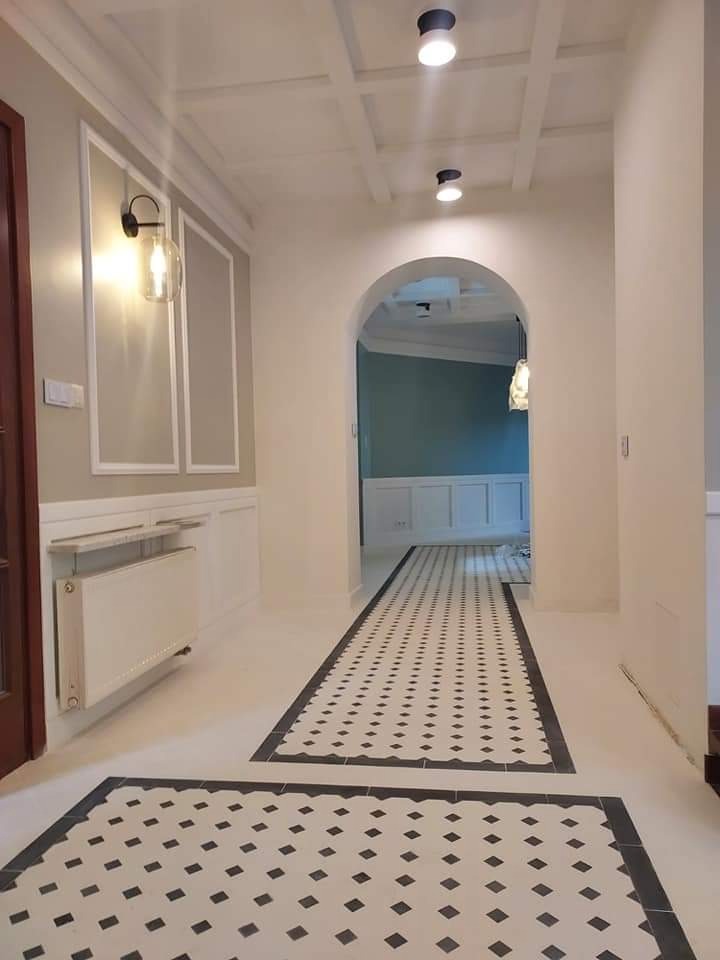 Wykończony korytarz w stylu klasycznym z białą boazerią, dekoracyjną listwą ścienną, grzejnikiem i wzorzystą podłogą z czarno-białych płytek, oświetlony nowoczesnymi lampami sufitowymi.