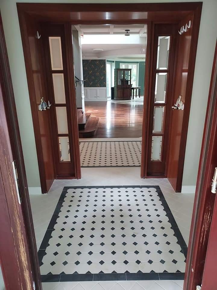 Korytarz z wzorzystą, czarno-białą podłogą z płytek ceramicznych, widok przez drewniane drzwi do salonu z parkietem i tapetą w kwiaty.