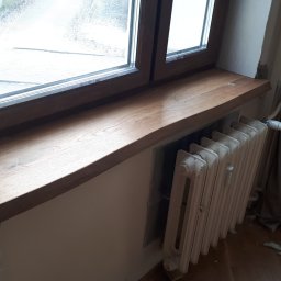 Groch-Bud - Montaż drewnianego parapetu okiennego, widoczne ślady pianki montażowej na ścianie i resztki na podłodze, grzejnik żeliwny pod oknem.