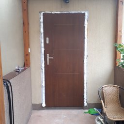 Groch-Bud - Brązowe drzwi wejściowe z ościeżnicą uszczelnianą pianką montażową, widok z zewnątrz, fragment tarasu z krzesłem i narzędziami w tle.