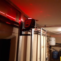 Groch-Bud - Laser krzyżowy wyznaczający poziom na ścianie z metalowych profili, widoczne czerwone linie lasera i elementy instalacji centralnego ogrzewania w tle.