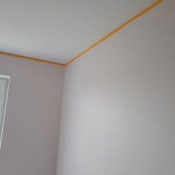 Groch-Bud - Świeżo pomalowane jasne ściany z żółtą taśmą malarską przy suficie w pomieszczeniu w Osieku nad Notecią, widoczne gniazdka elektryczne.