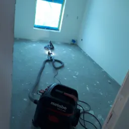 Pokój w trakcie remontu, z gładziami na ścianach i podłodze, widoczny odkurzacz przemysłowy Metabo ASR 35 ACP oraz szlifierka do gładzi na wysięgniku, okno zabezpieczone folią.