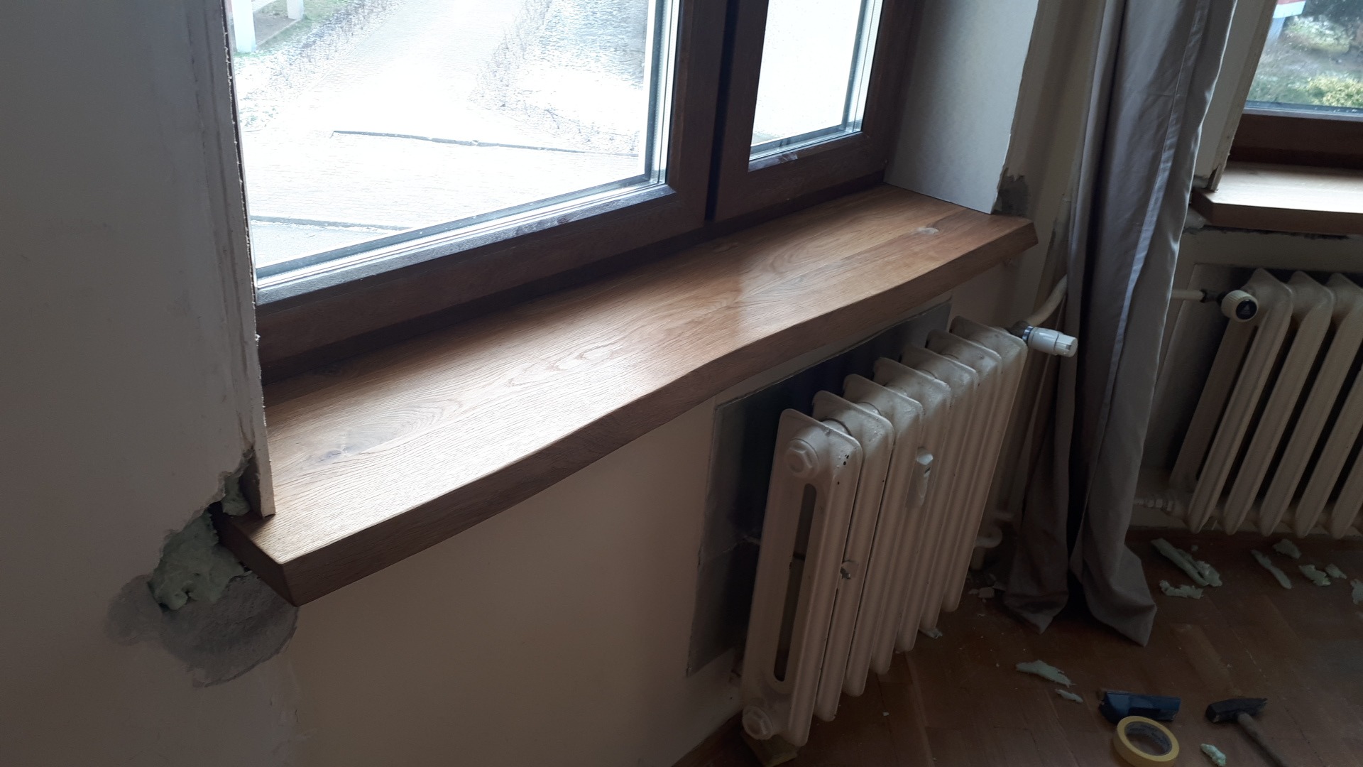 Montaż drewnianego parapetu okiennego, widoczne ślady pianki montażowej na ścianie i resztki na podłodze, grzejnik żeliwny pod oknem.