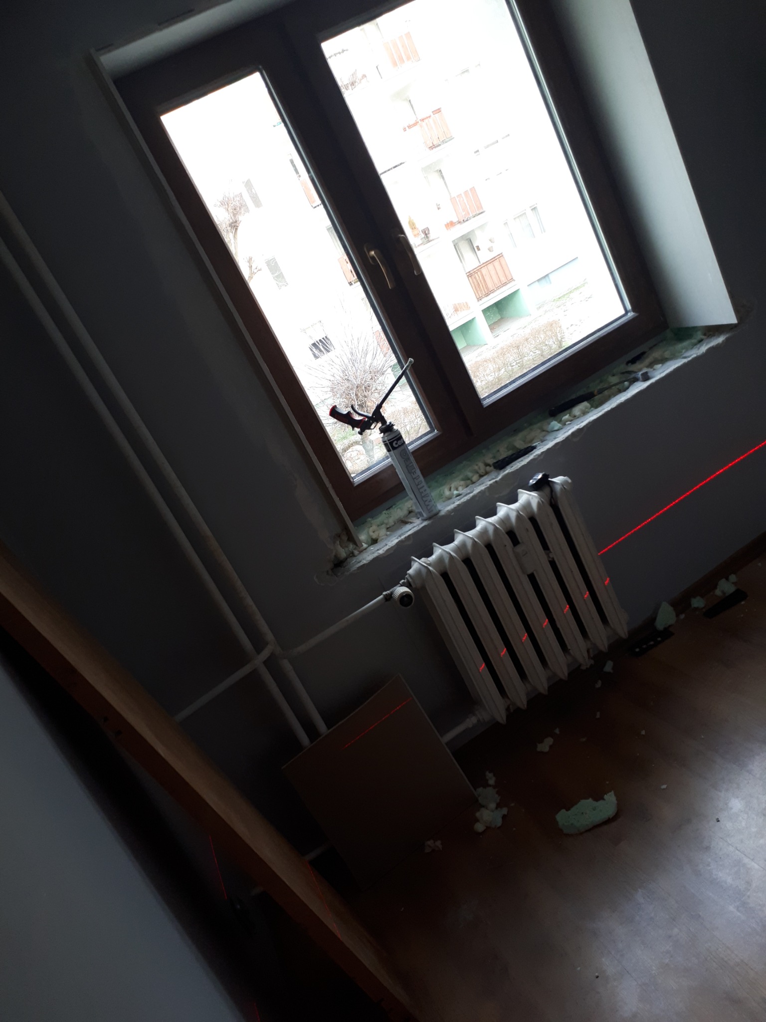 Montaż okna z użyciem pianki montażowej, widoczny grzejnik, poziomica laserowa i fragment podłogi z resztkami pianki.