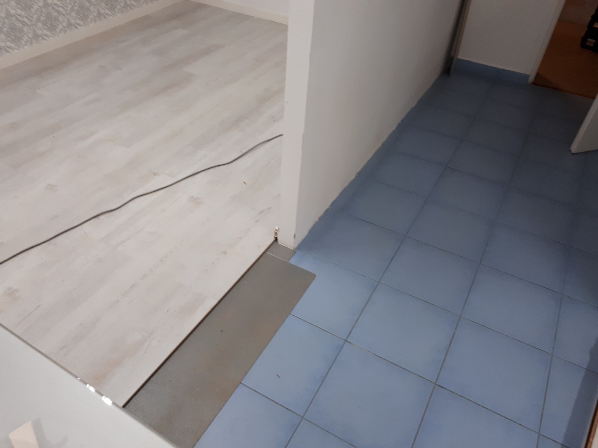 Połączenie dwóch rodzajów podłóg: panele podłogowe w odcieniach bieli i niebieskie płytki ceramiczne, oddzielone szarą listwą przejściową, widoczne kable elektryczne na panelach.