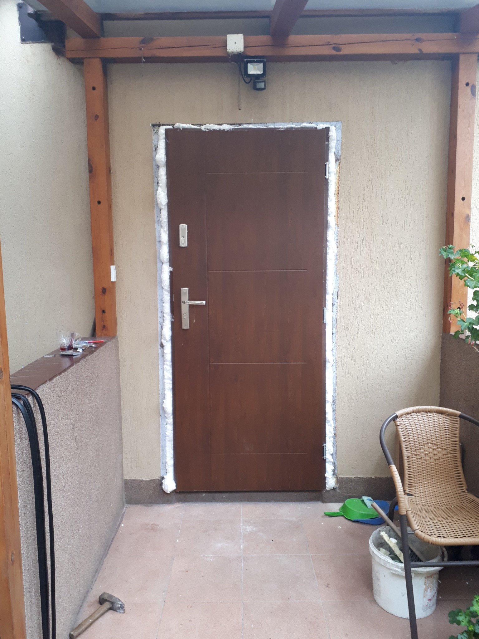 Brązowe drzwi wejściowe z ościeżnicą uszczelnianą pianką montażową, widok z zewnątrz, fragment tarasu z krzesłem i narzędziami w tle.