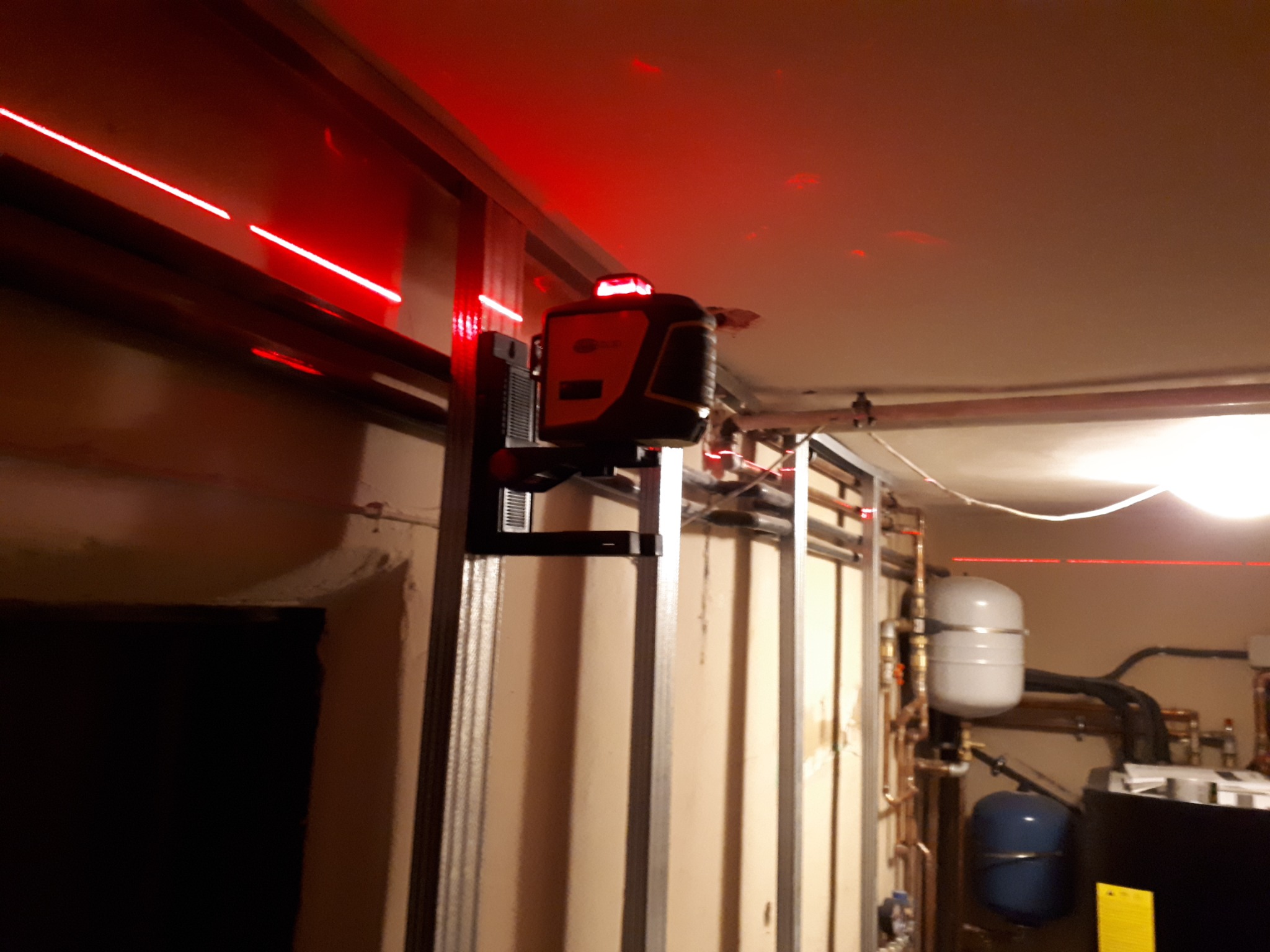 Laser krzyżowy wyznaczający poziom na ścianie z metalowych profili, widoczne czerwone linie lasera i elementy instalacji centralnego ogrzewania w tle.