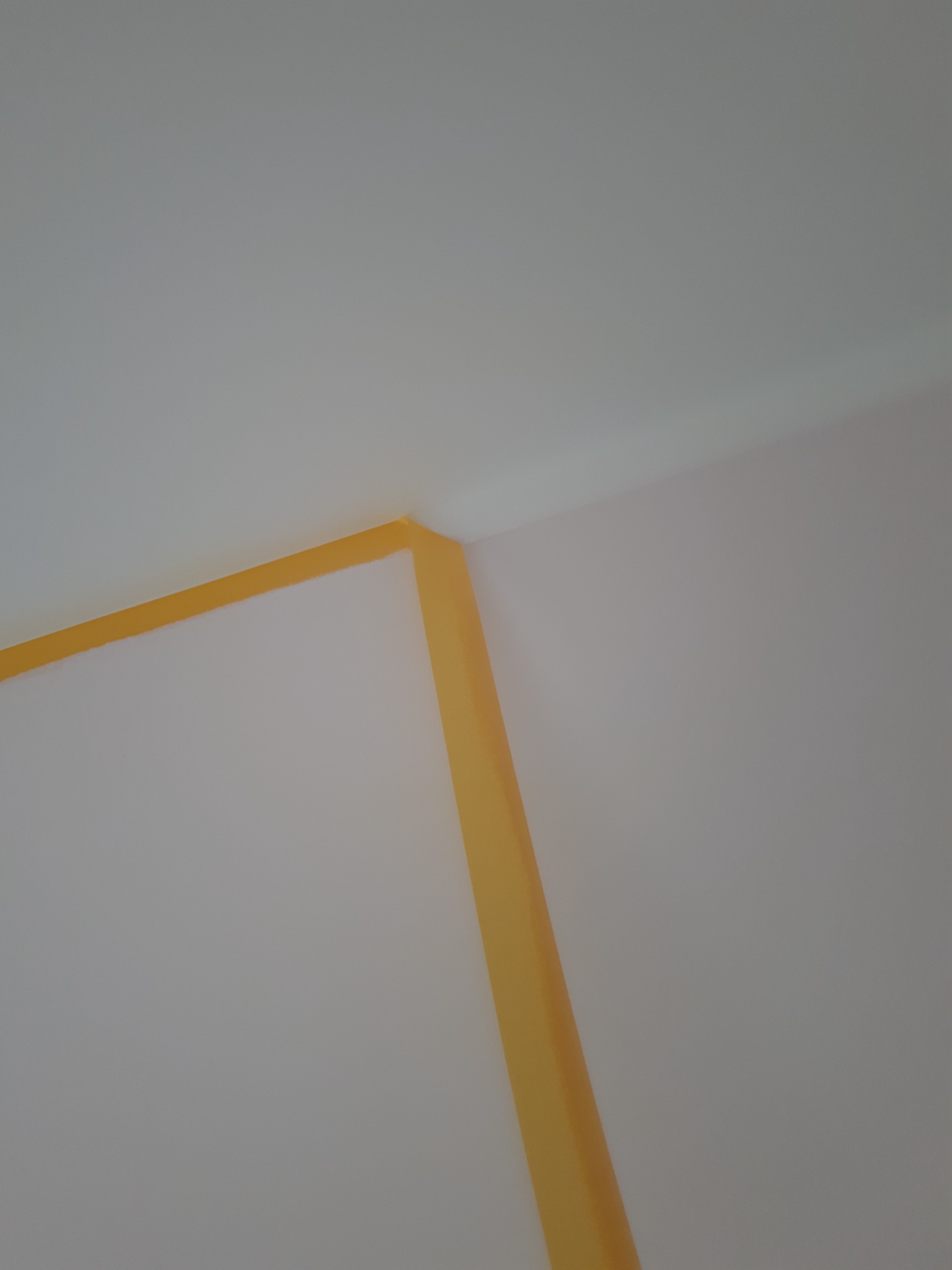 Żółta taśma malarska zabezpieczająca narożnik ściany i sufit przed malowaniem, widok z dołu pod kątem.