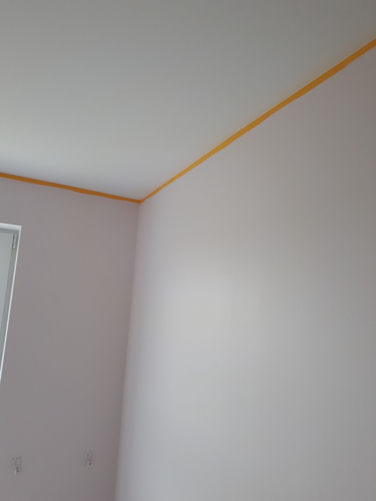 Świeżo pomalowane jasne ściany z żółtą taśmą malarską przy suficie w pomieszczeniu w Osieku nad Notecią, widoczne gniazdka elektryczne.