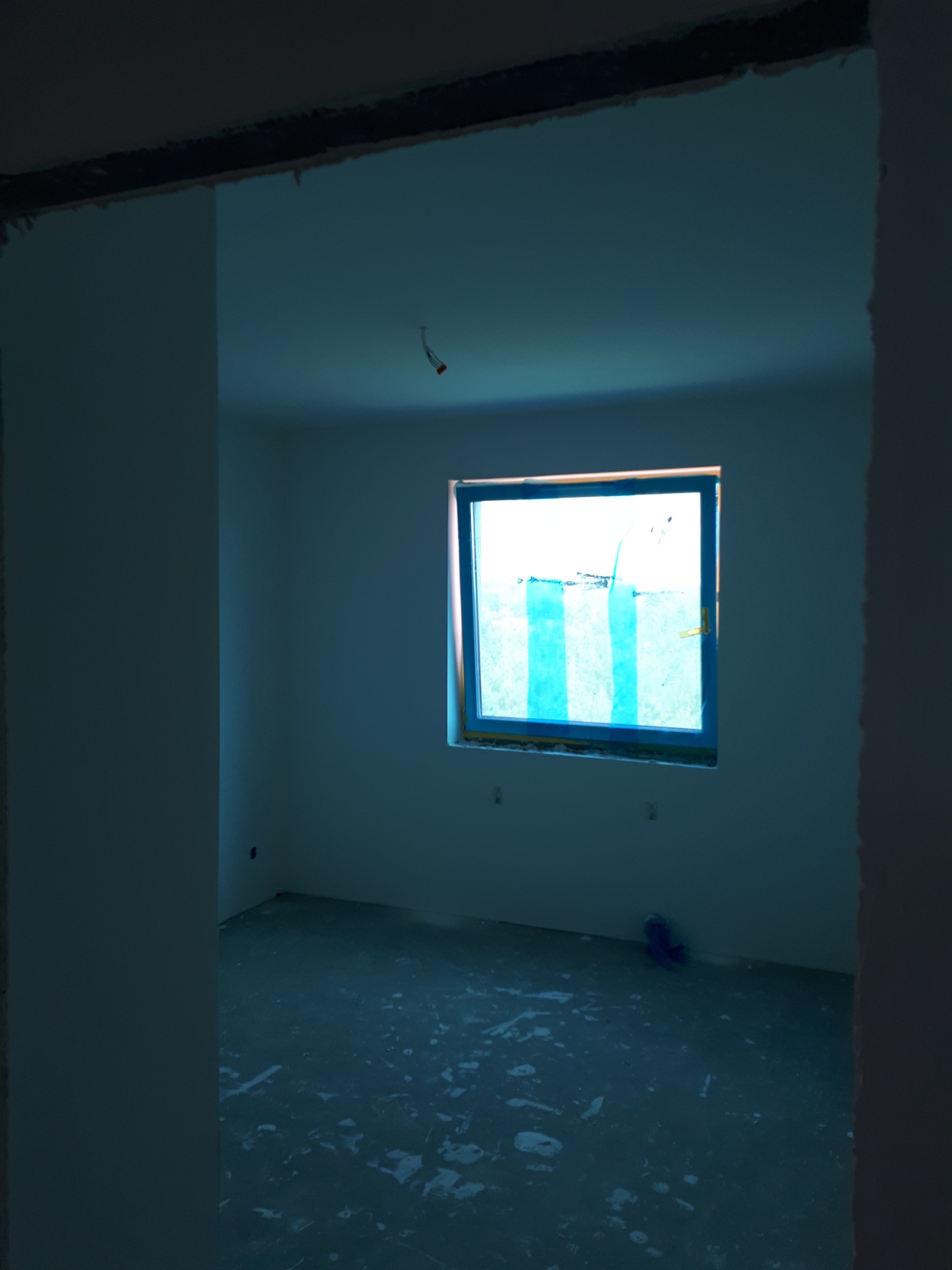 Pokój w trakcie prac wykończeniowych, widoczne okno zabezpieczone niebieską folią malarską, ślady zaprawy na podłodze, surowy stan ścian przed malowaniem.