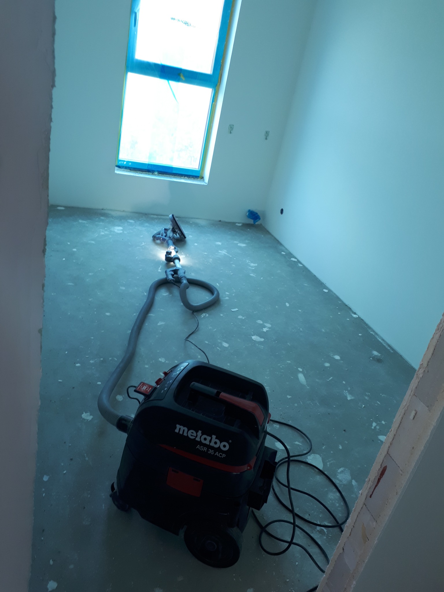 Pokój w trakcie remontu, z gładziami na ścianach i podłodze, widoczny odkurzacz przemysłowy Metabo ASR 35 ACP oraz szlifierka do gładzi na wysięgniku, okno zabezpieczone folią.
