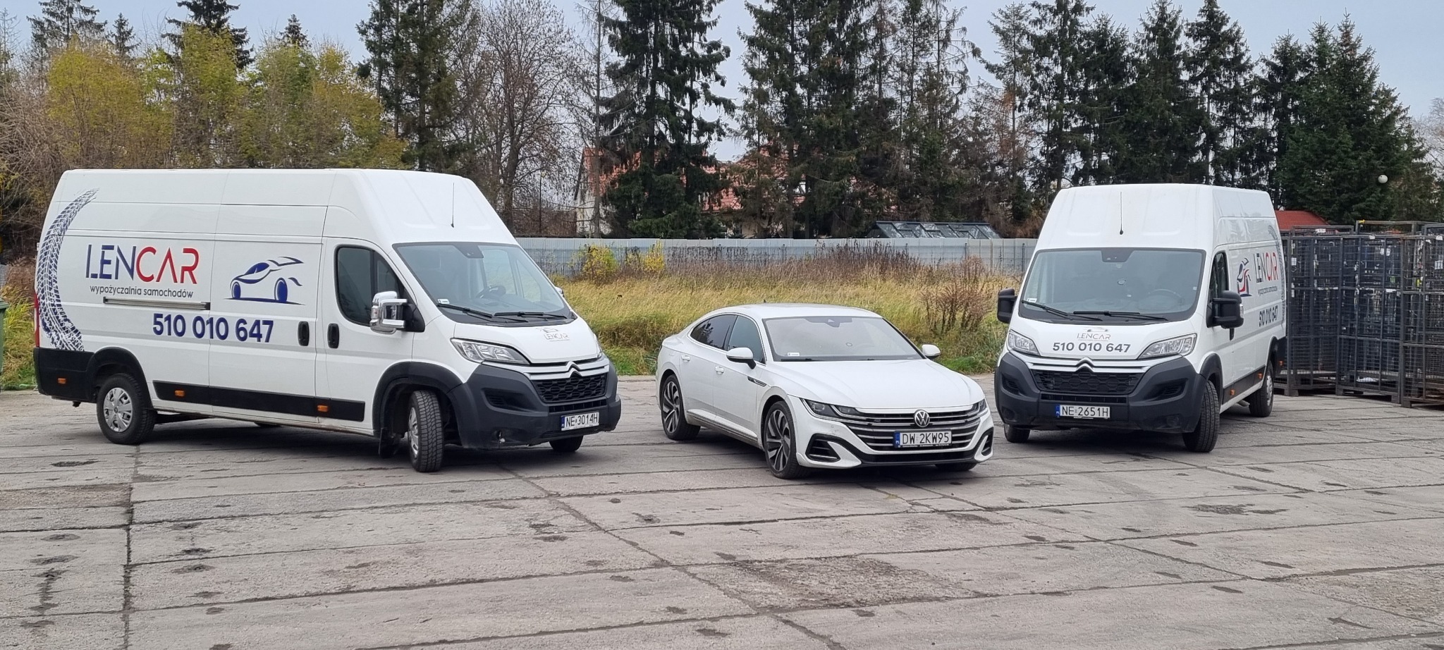 Dwa białe busy dostawcze z logo wypożyczalni samochodów Lencar oraz biały samochód osobowy Volkswagen Arteon na betonowym placu w pochmurny dzień.