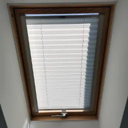 Okno dachowe z drewnianą ramą i plisowaną roletą w kolorze białym, zamontowane w skosie dachu, widoczne od wewnątrz, z delikatnym oświetleniem z boku.