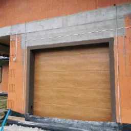 Montaż segmentowej bramy garażowej z imitacją drewna w nowym budynku z czerwonej cegły, widoczne kable i folia fundamentowa.