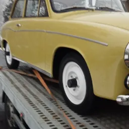 Żółty zabytkowy samochód zabezpieczony pasami transportowymi, umieszczony na platformie lawety w pochmurny dzień.