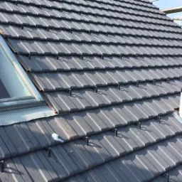 Szare dachówki ceramiczne z zamkami na skośnym dachu, widoczne okno dachowe z metalową obróbką, kominek wentylacyjny i pracownik na rusztowaniu w tle.