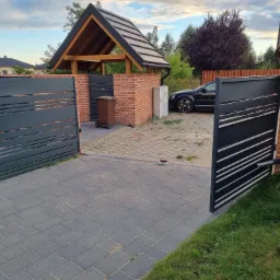 Nowoczesna, automatyczna brama wjazdowa z poziomymi panelami, zamontowana między murowanymi słupkami z czerwonej cegły, z widocznym napędem i kostką brukową na podjeździe.