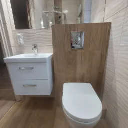 Nowoczesna łazienka po remoncie: biała umywalka z szafką, toaleta z drewnianą obudową spłuczki i beżowe płytki imitujące drewno na ścianach i podłodze.