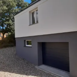 Dwukolorowa elewacja domu z garażem, górna część biała, dolna szara, widok z zewnątrz na podjazd z kamyków i drzewa w tle.