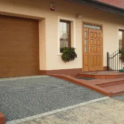 Podjazd do garażu i wejście do domu wyłożone szarą, ażurową eco-kratką trawnikowo-parkingową Pebart, obrzeża z czerwonej cegły klinkierowej.