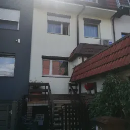 Dwukondygnacyjny dom z jasną elewacją i roletami zewnętrznymi, widoczny z zewnątrz, z fragmentem ciemnoszarej ściany sąsiedniego budynku po lewej stronie, czerwonym dachem i zielenią w ogrodzie.